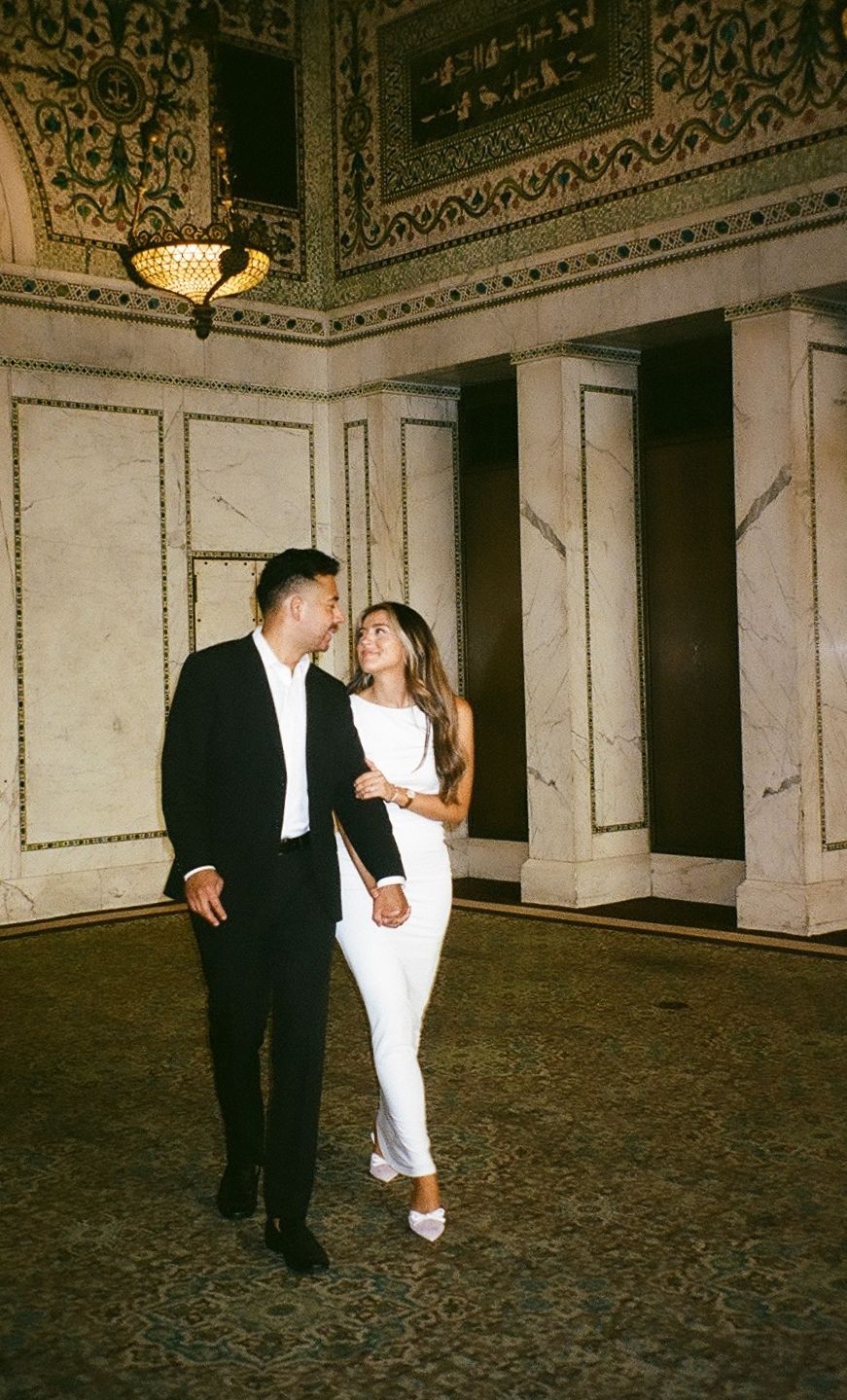 Couple dancing together in elegant marble ballroom with ornate ceiling and vintage chandeliers.