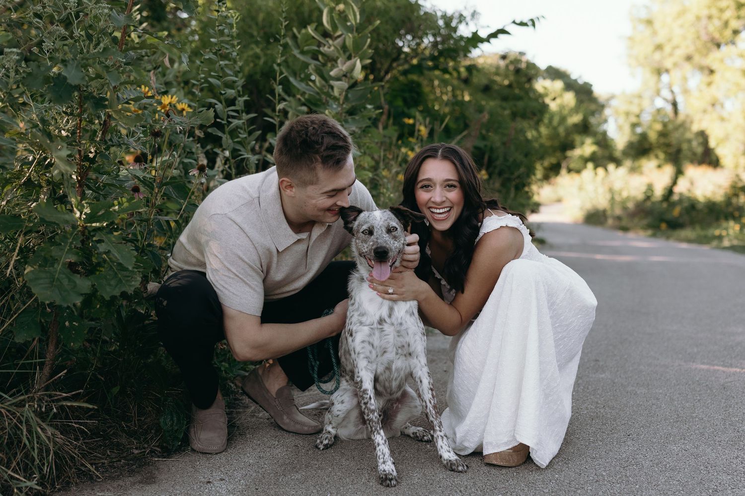 A dog with spotted fur poses on a paved path with two people crouching beside it in a white dress and tan outfit.
