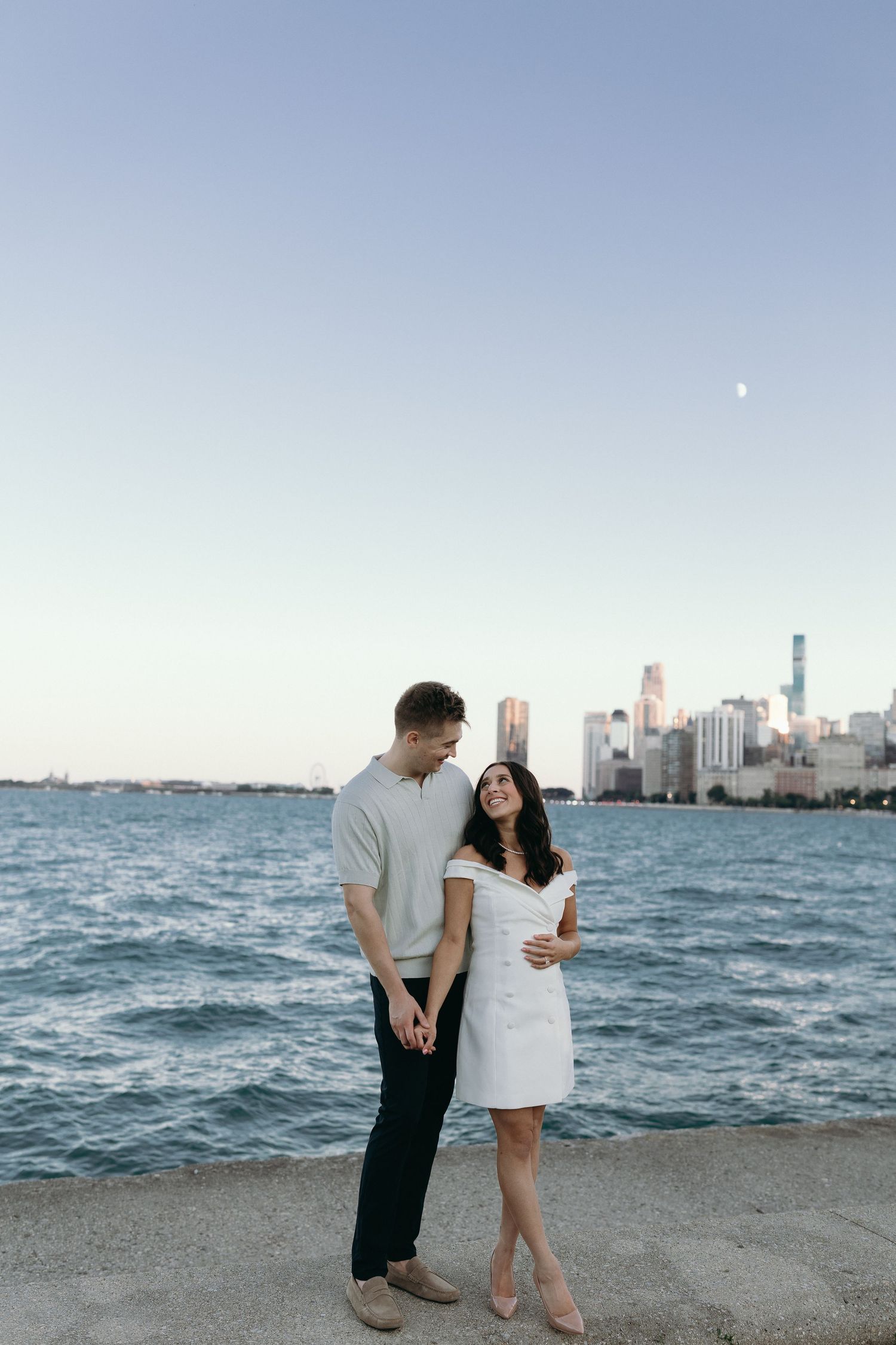 A couple stands together on the shore of Lake Michigan with Chicago's skyline visible at dusk and moon in the sky.