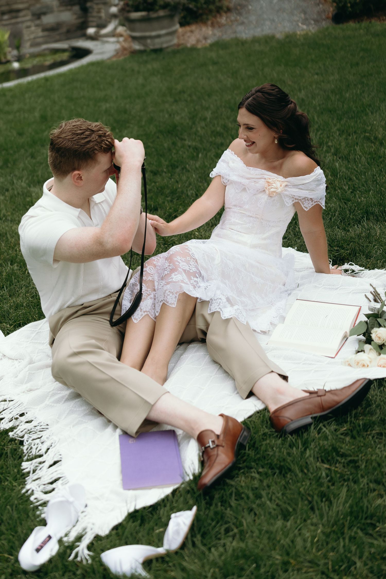 A playful wedding couple resting on green grass wearing a white gown and beige suit during an outdoor photoshoot.