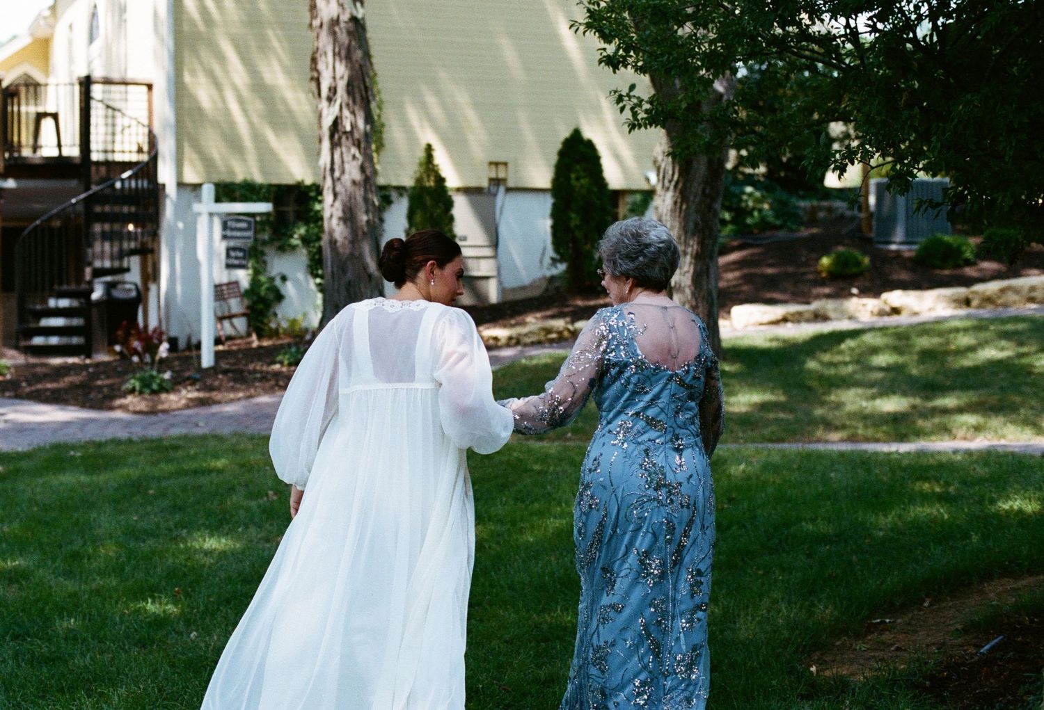 Two women in elegant dresses hold hands walking through a lush green garden near a white house. on film