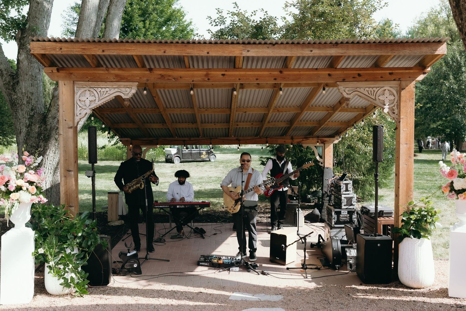 Live wedding band performing under rustic wooden pergola with corrugated metal roof at outdoor venue.