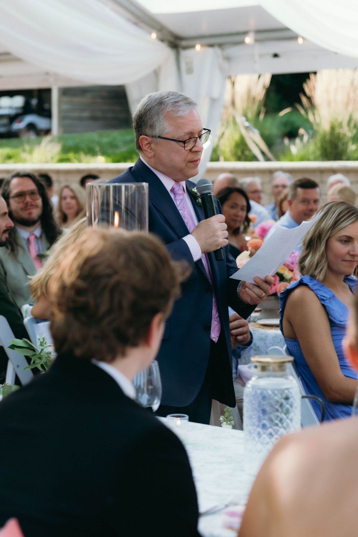 Older gentleman in navy suit giving wedding speech into microphone before seated reception guests.