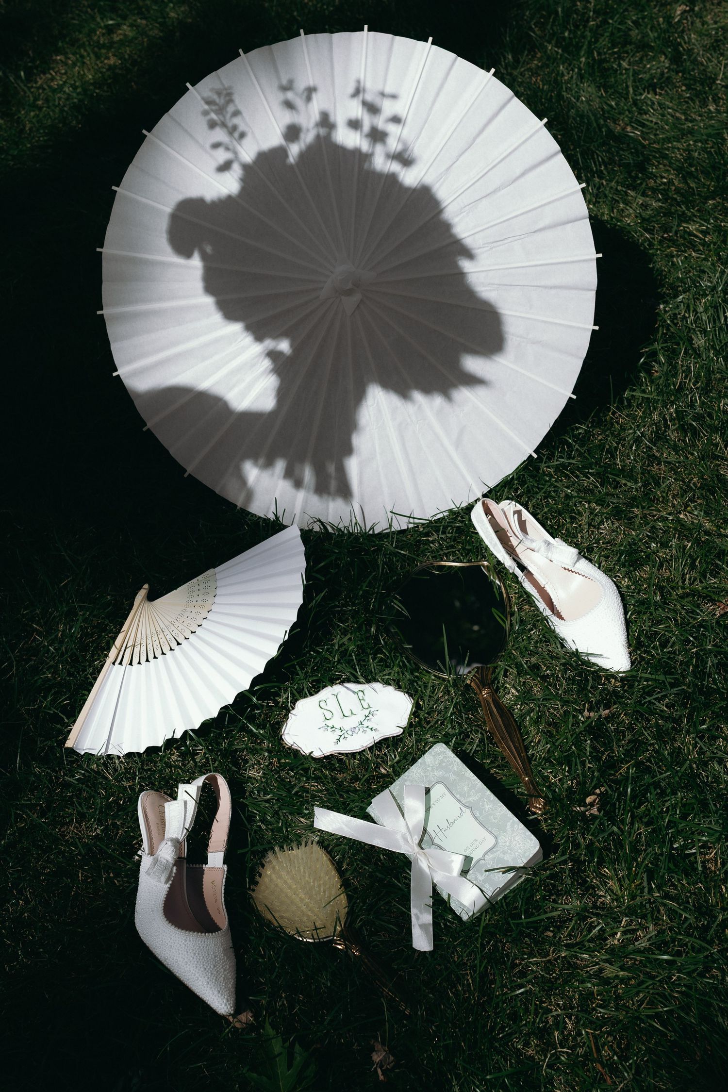 Bridal accessories flatlay on grass featuring white parasol, fan, heels, sunglasses, and monogrammed items.