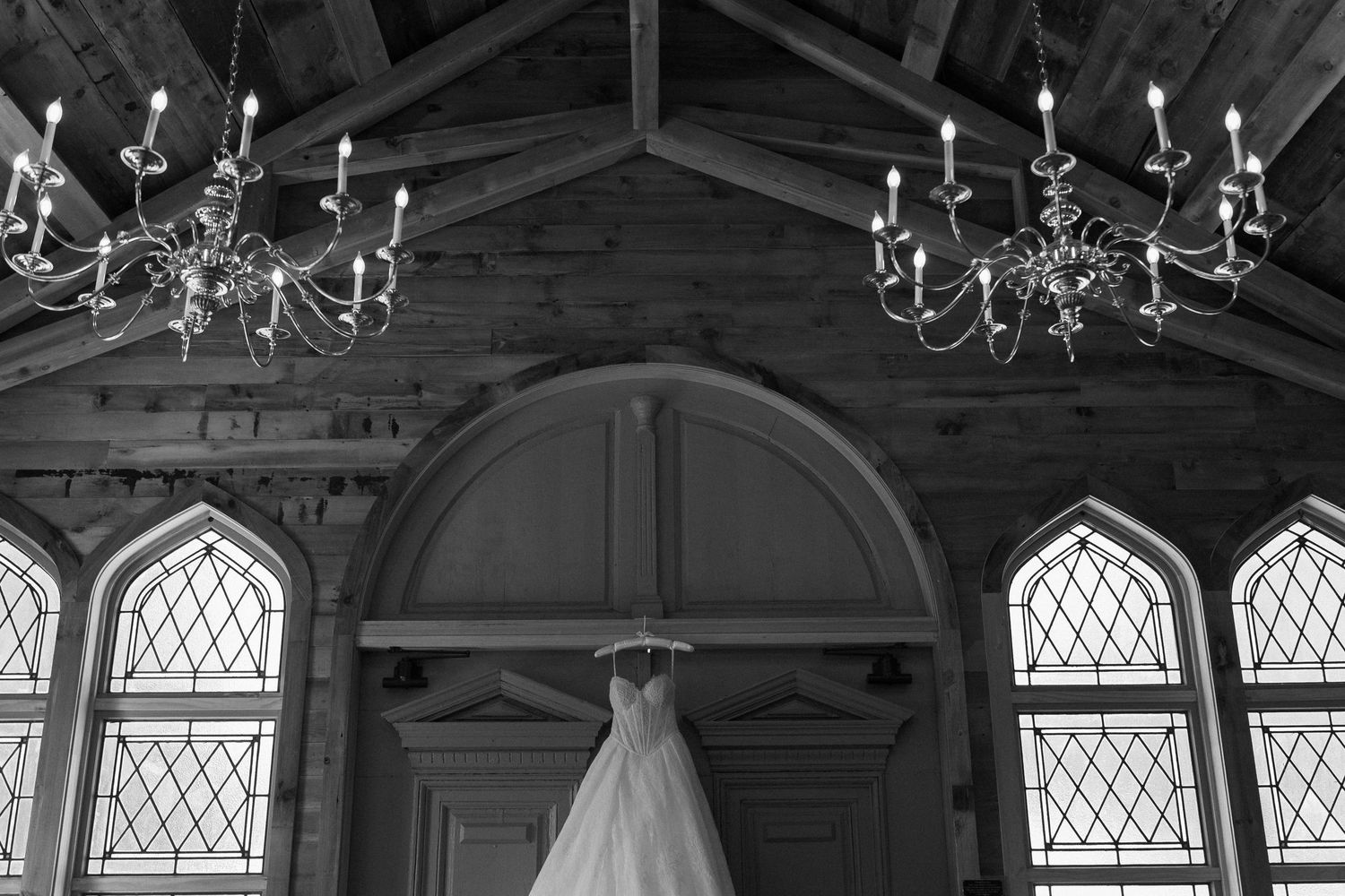 Black and white photo of a wedding dress hanging in a rustic chapel with arched windows and elegant chandeliers.