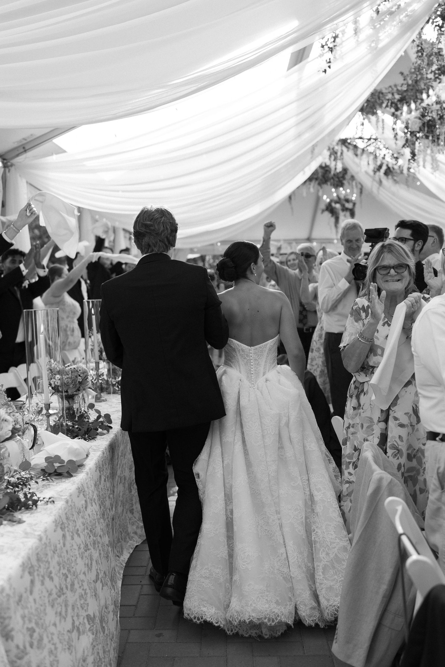 Black and white photo of bride and groom entering wedding reception as guests celebrate.