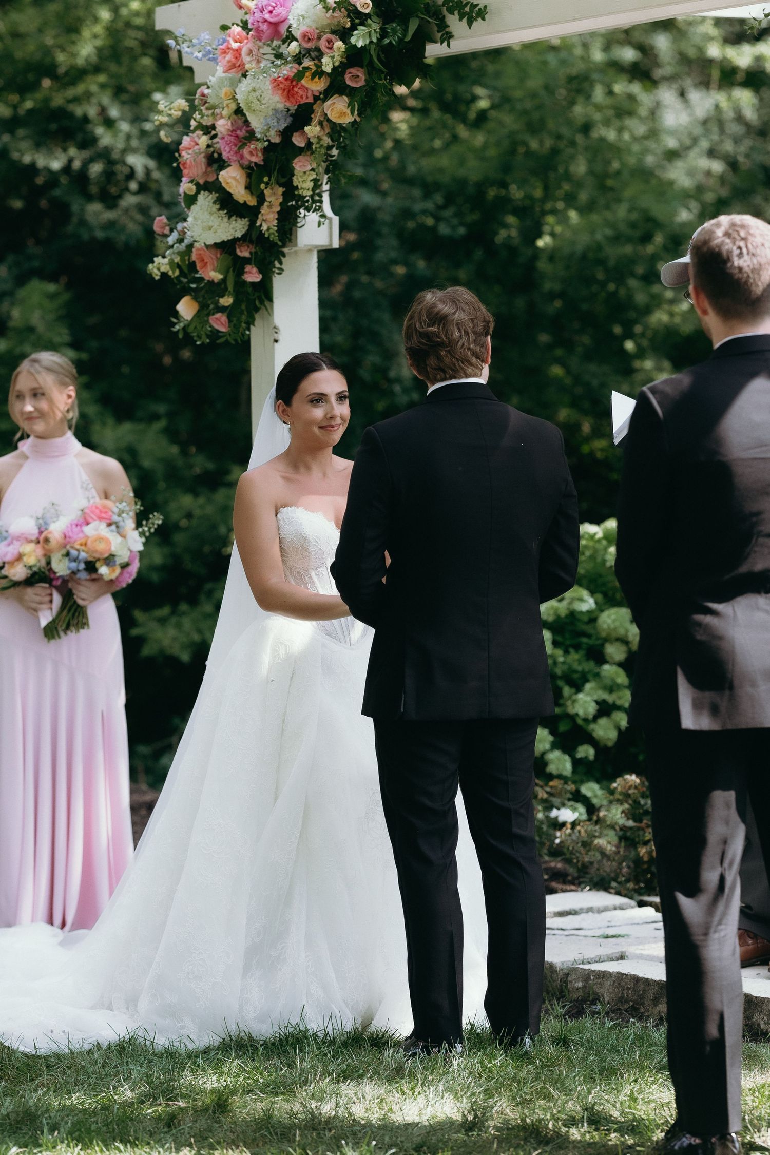 Bride and groom exchange vows outdoors under floral arch, with bridesmaid in pink dress holding bouquet nearby.