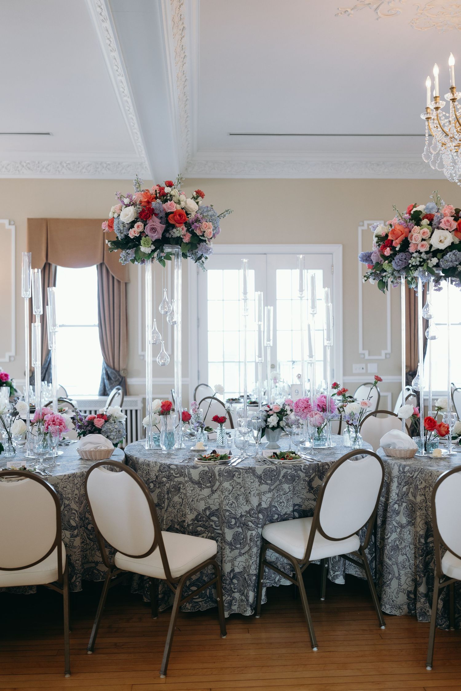 Elegant wedding reception table with white chairs, floral centerpieces, and grey patterned tablecloth running along windows.