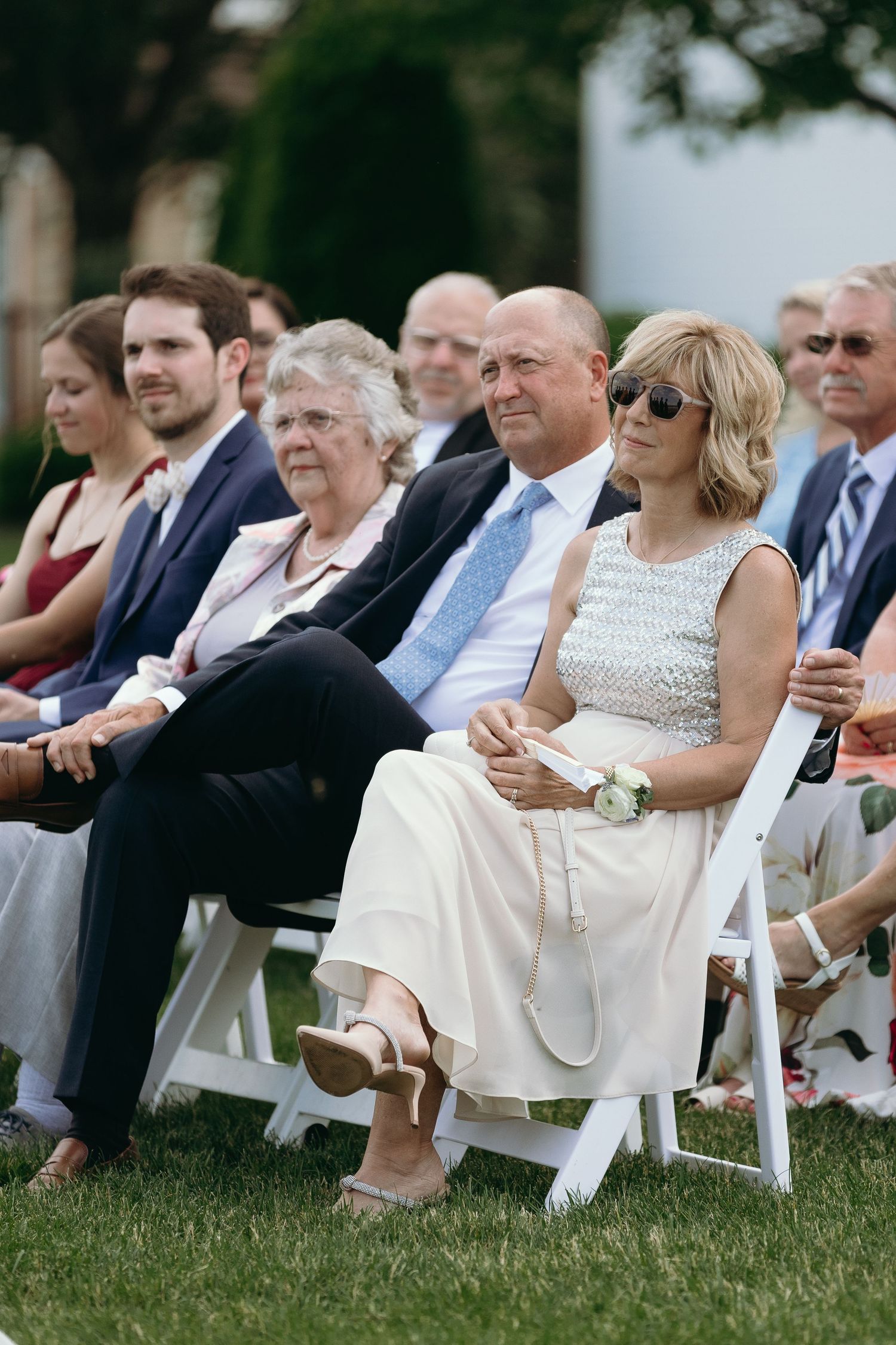 Wedding guests seated in white chairs on a lawn, with some wearing sunglasses and formal attire at an outdoor ceremony.