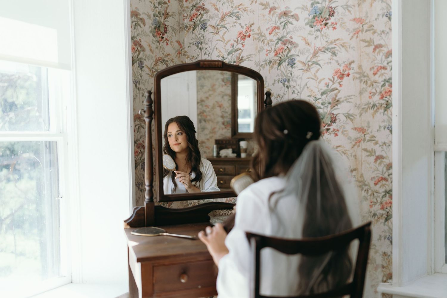 A woman getting ready at a vintage vanity with floral wallpaper in the background.