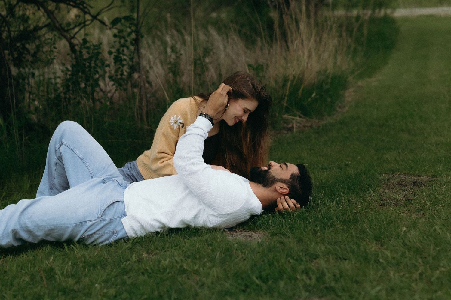 Romantic couple lying on green grass outdoors, woman leaning over man in tender moment, casual attire, park setting.