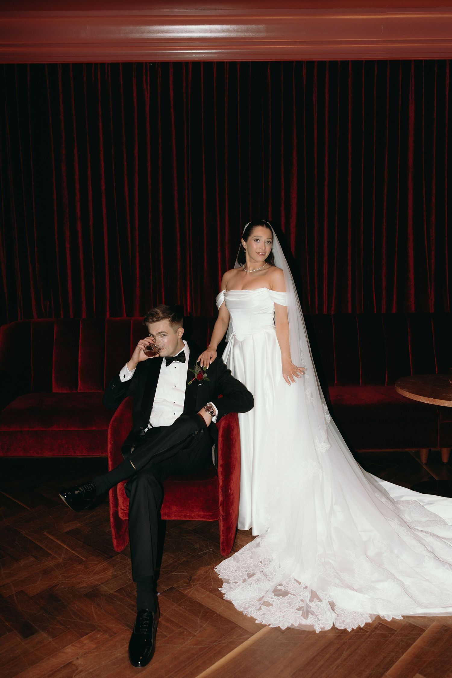Bride in white off-shoulder gown with veil stands behind groom in black tuxedo seated on red velvet chair.