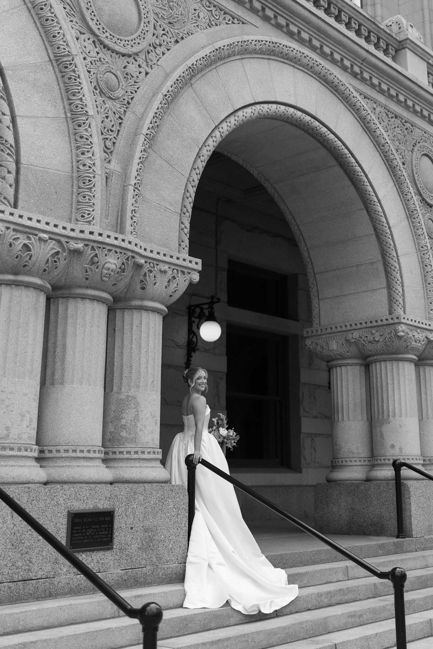 A bride in a white wedding gown poses elegantly along an ornate stone colonnade with arched architecture.