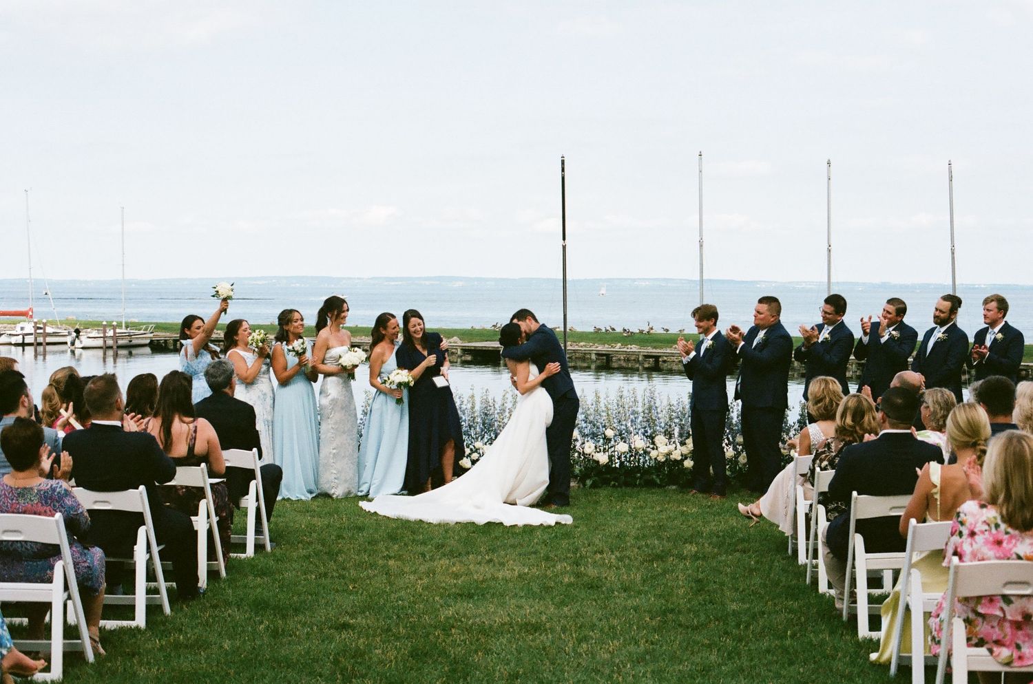 Outdoor waterfront wedding ceremony with bride and groom kissing, bridesmaids in blue, groomsmen in black suits.