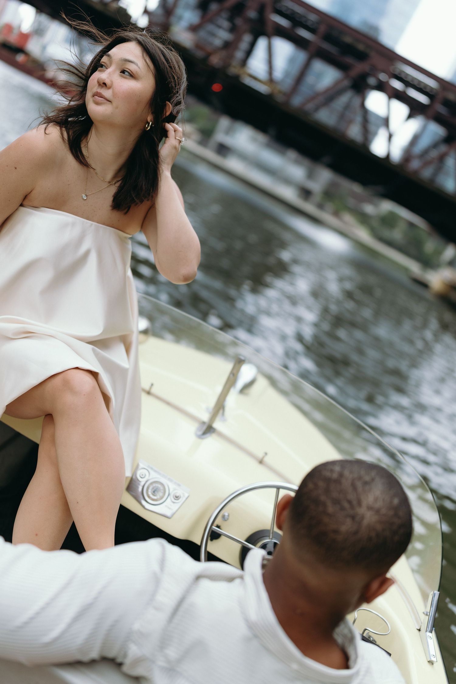 A woman in a white dress sits on a vintage boat with a man at the helm, with a city bridge in the background.