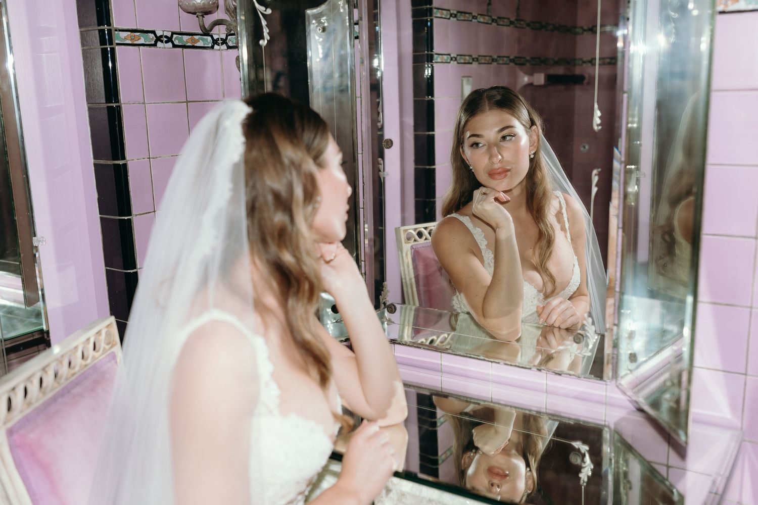 Reflection of person in wedding attire applying makeup in pink-tiled bathroom mirror.