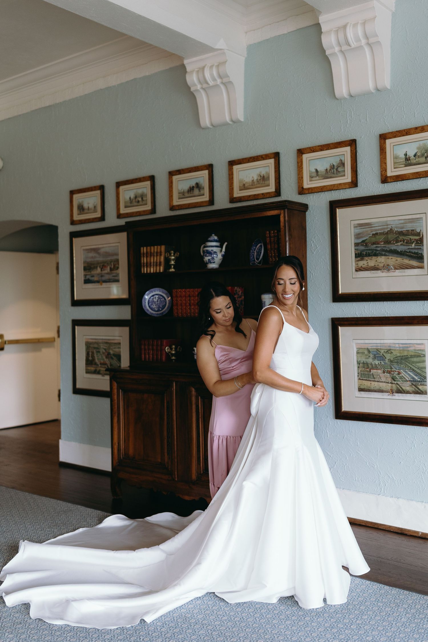 White wedding dress with long train being adjusted in a room with framed artwork.