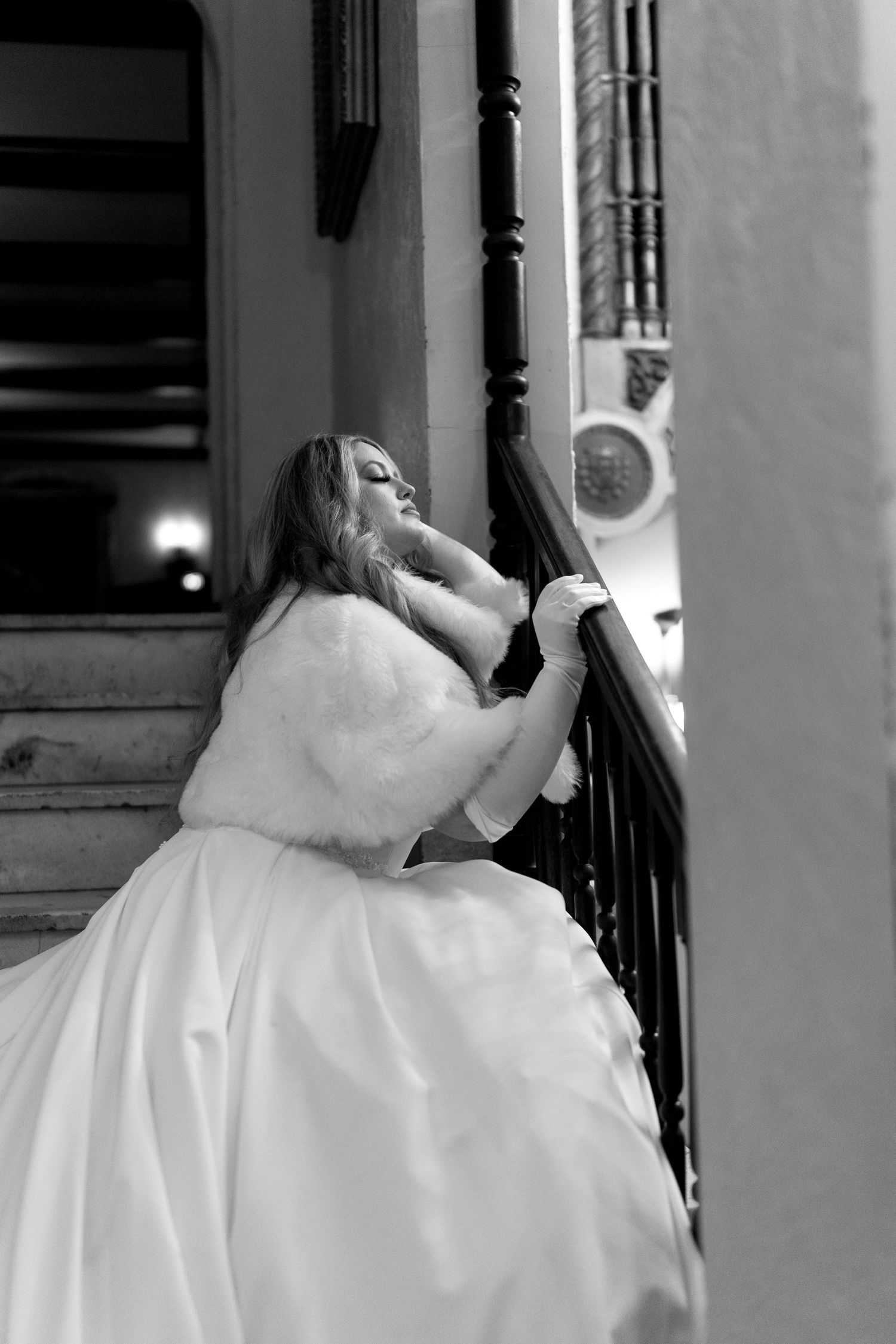 Bride in flowing white dress and fur wrap poses dramatically on grand staircase.