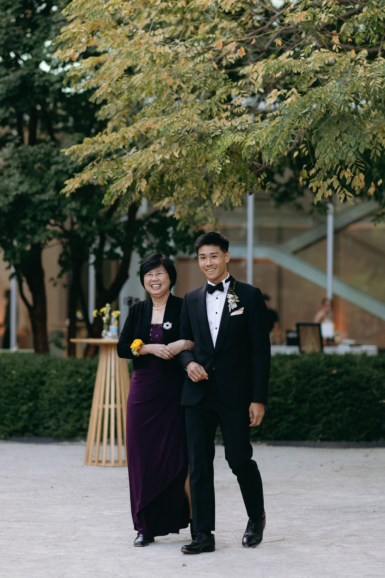 Wedding party members in formal attire walking together outdoors beneath green trees.