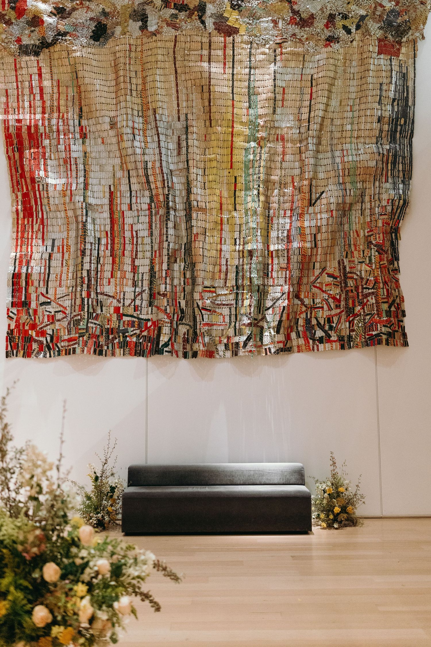 A decorative fabric installation with red and brown tones hangs above a black bench with floral arrangements.