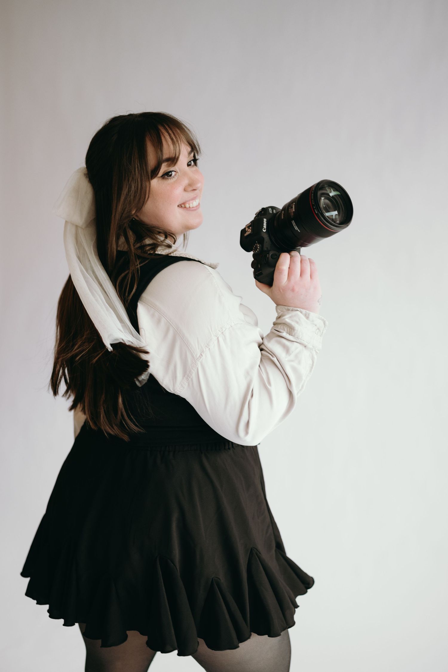 A photographer in a white top and black skirt holds a professional camera while smiling.