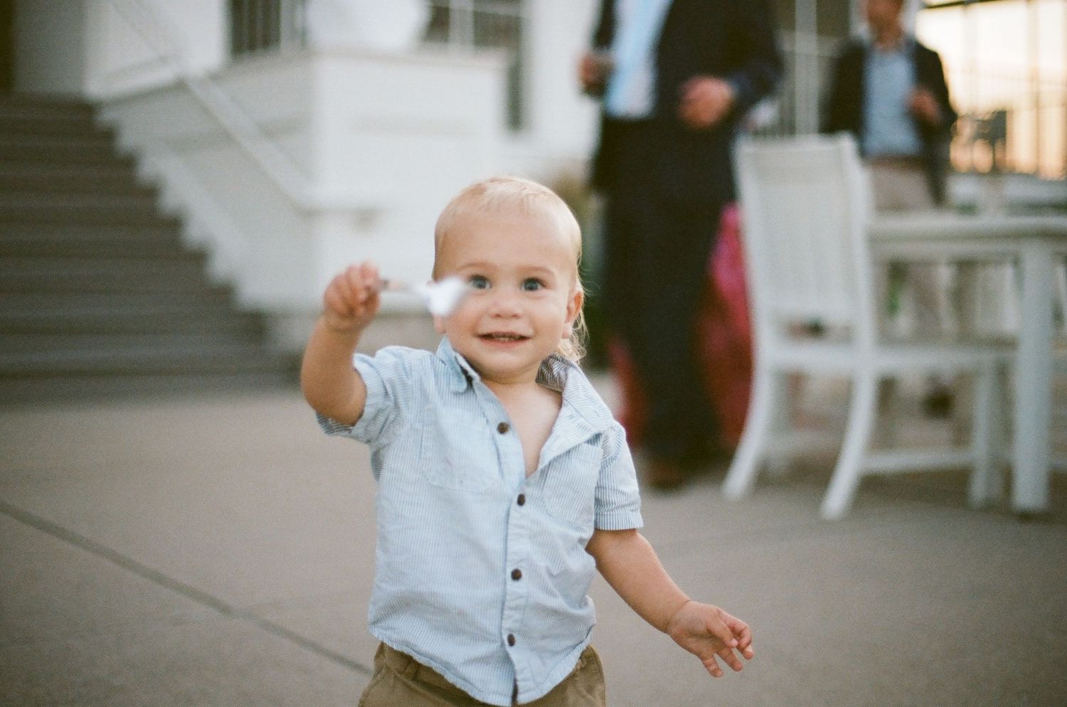A small child in a blue button-down shirt waves while standing on a porch in warm lighting.