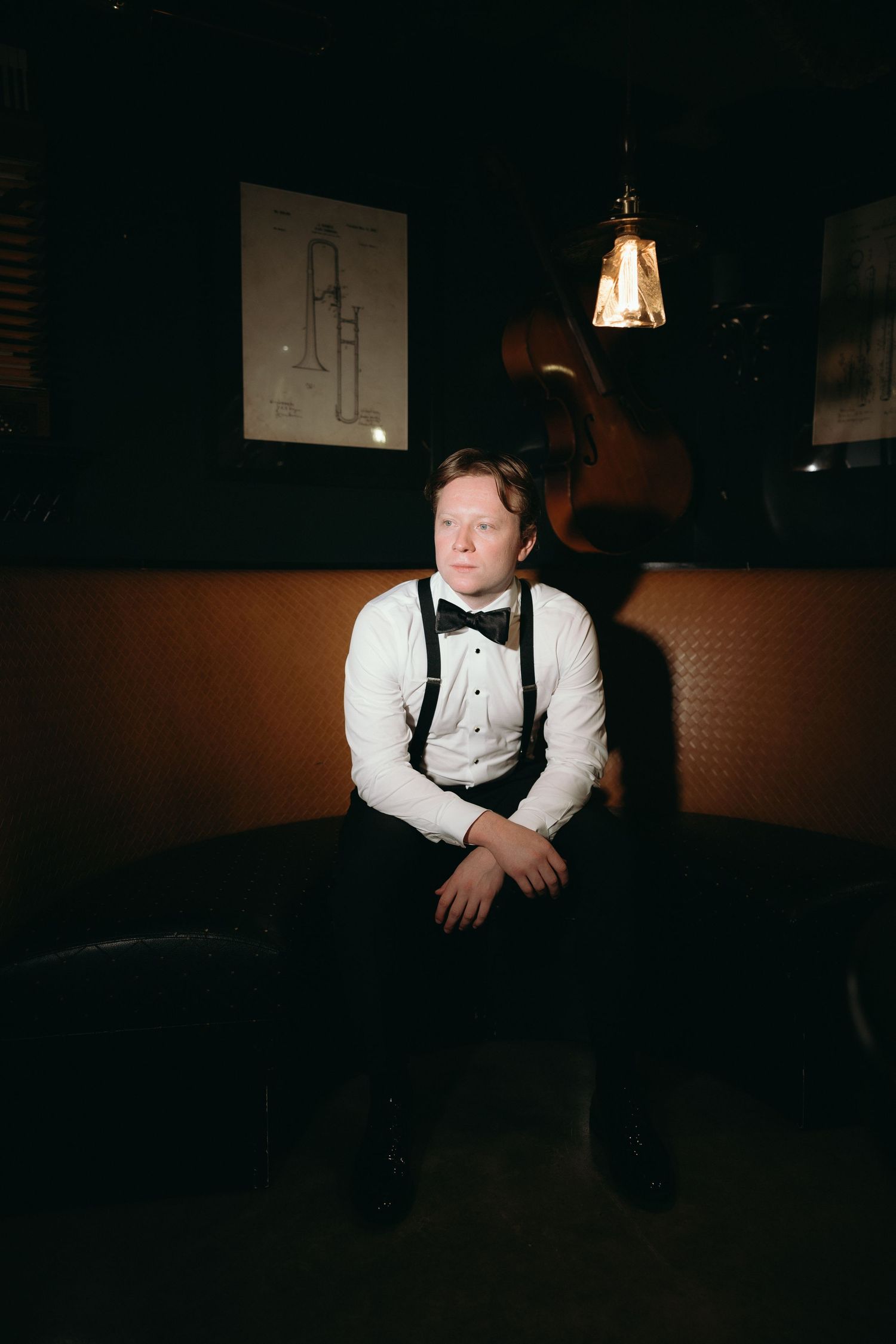 Young man in white shirt, bow tie, and suspenders sits in a dimly lit jazz bar with vintage decor and hanging light.