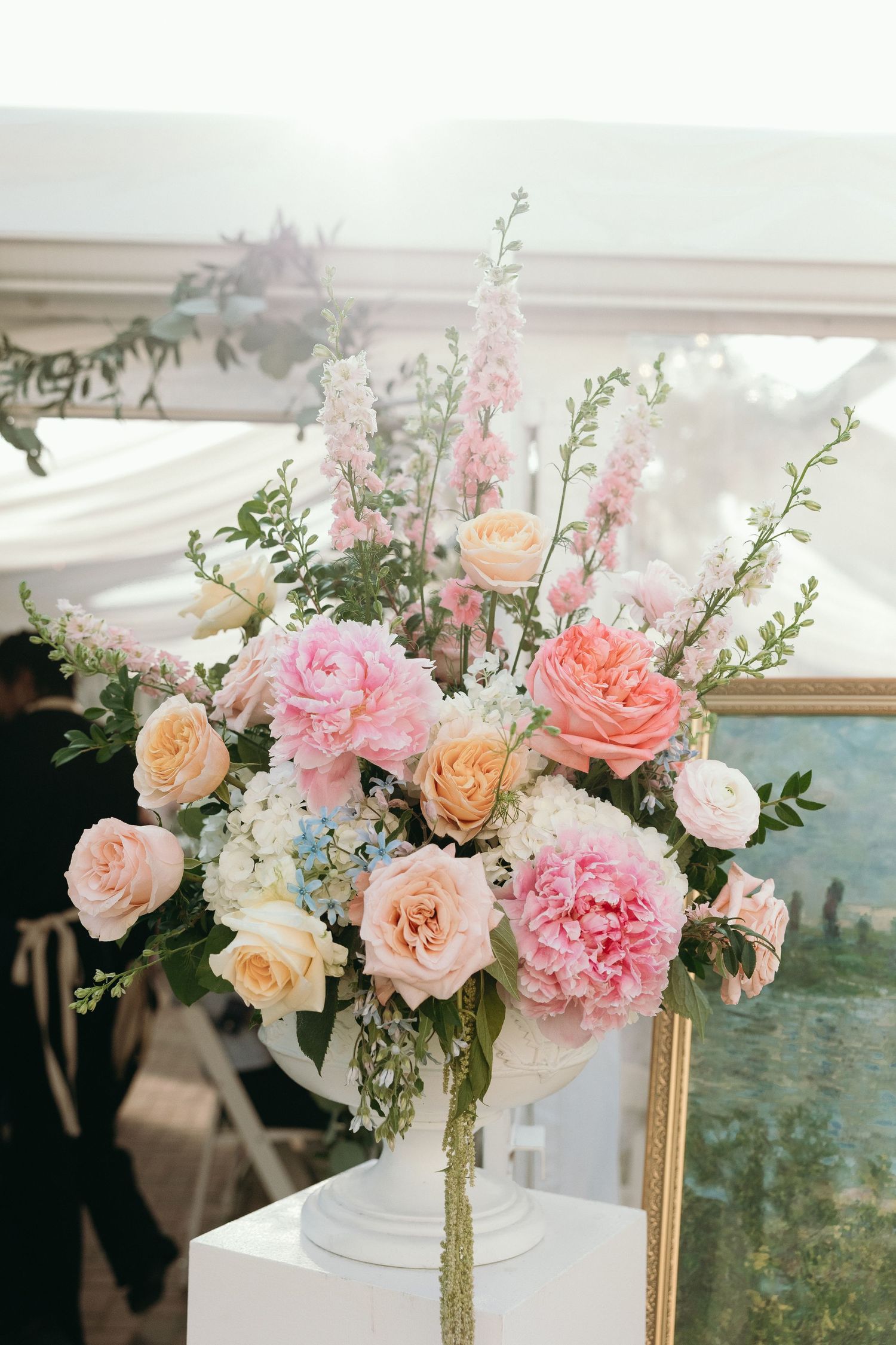 Elegant wedding floral arrangement featuring pink peonies, peach roses, and white hydrangeas in a white urn vase.