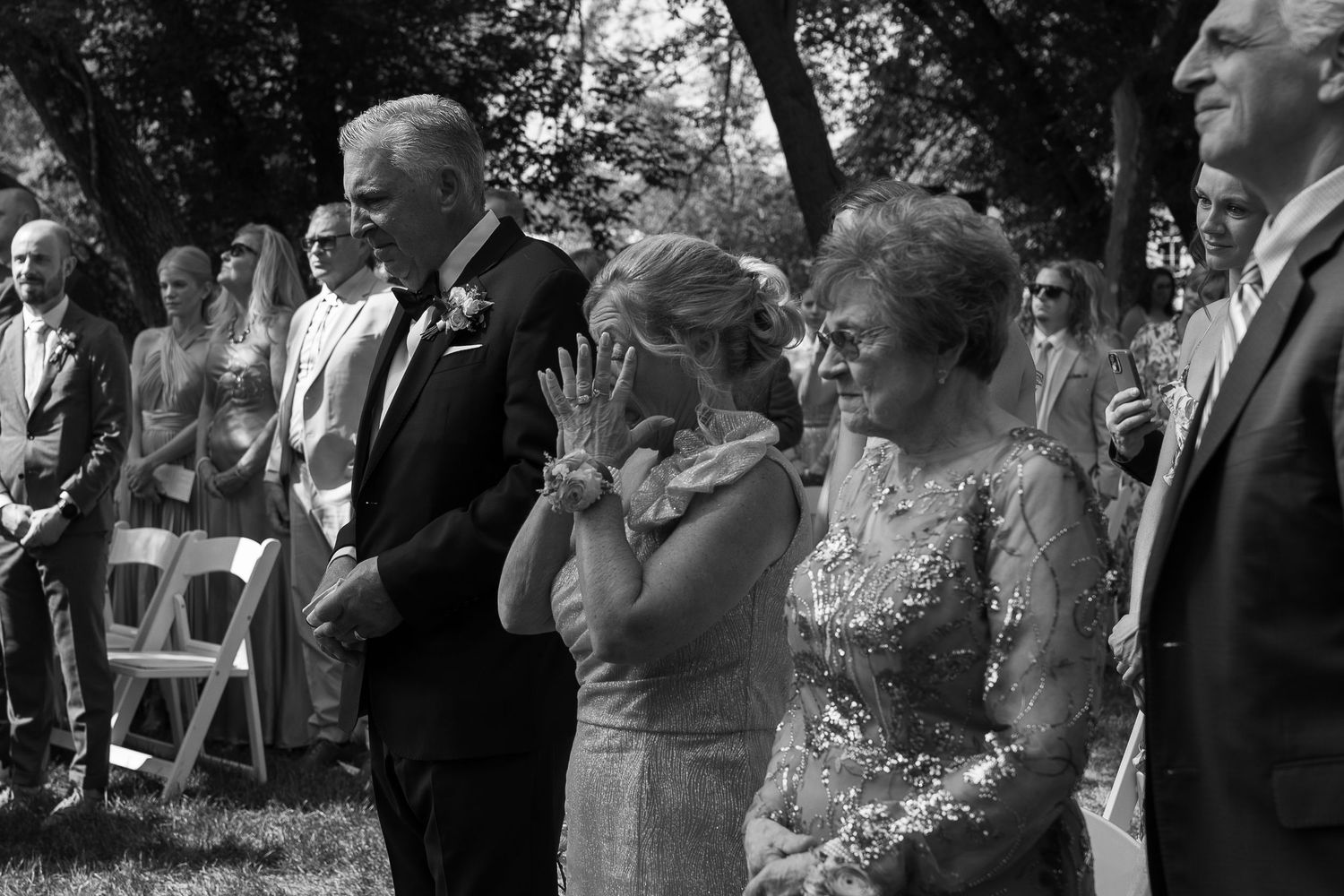 Black and white outdoor wedding ceremony with emotional guests, woman wiping tears, elegant attire, trees in background.