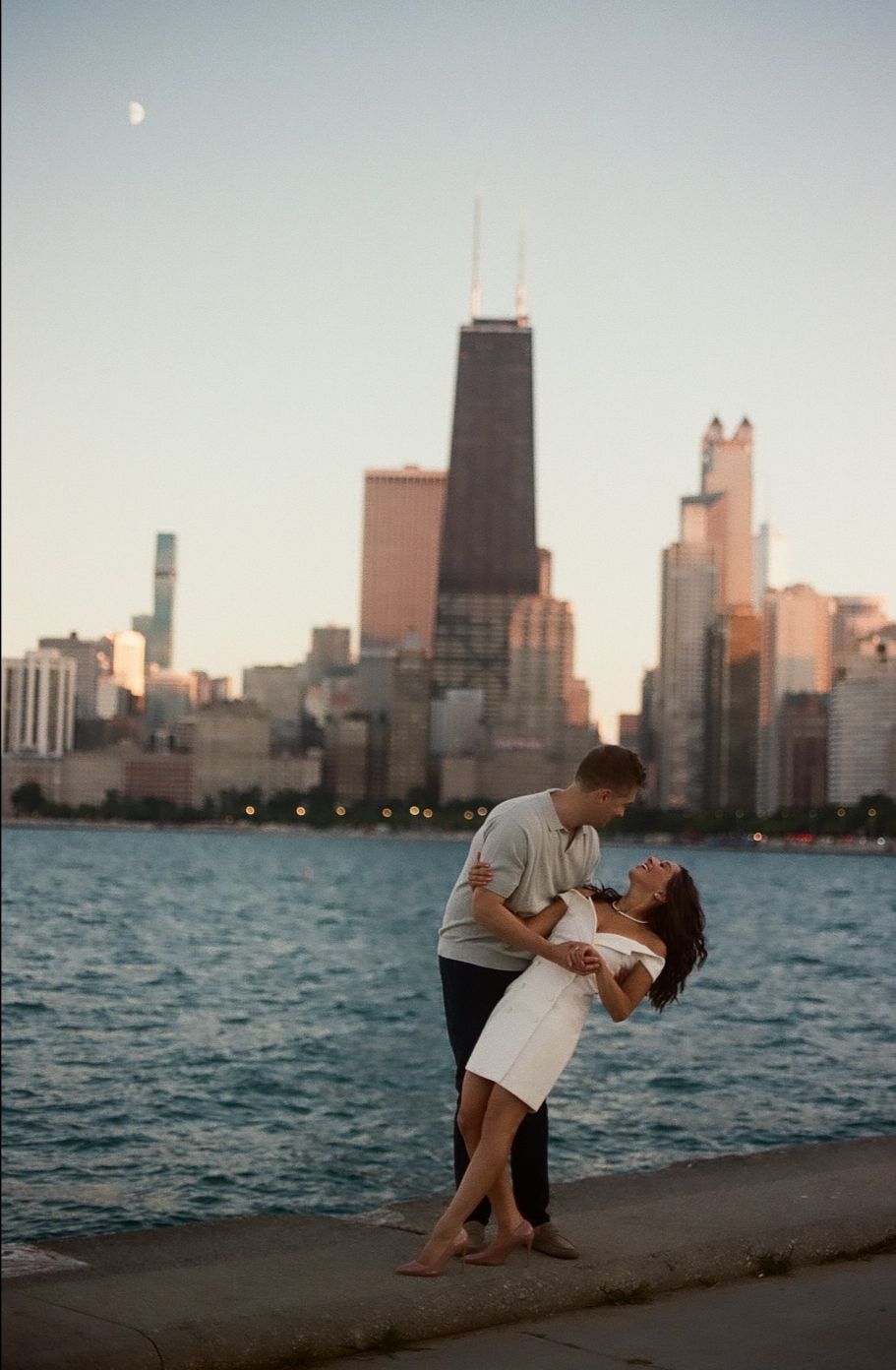 A romantic couple embraces on the Chicago lakefront at sunset with the iconic downtown skyline visible in the background.