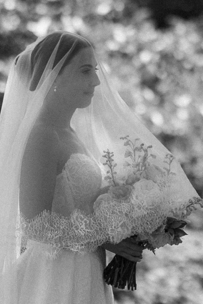 Black and white bridal portrait of a bride in lace gown and veil holding a floral bouquet.