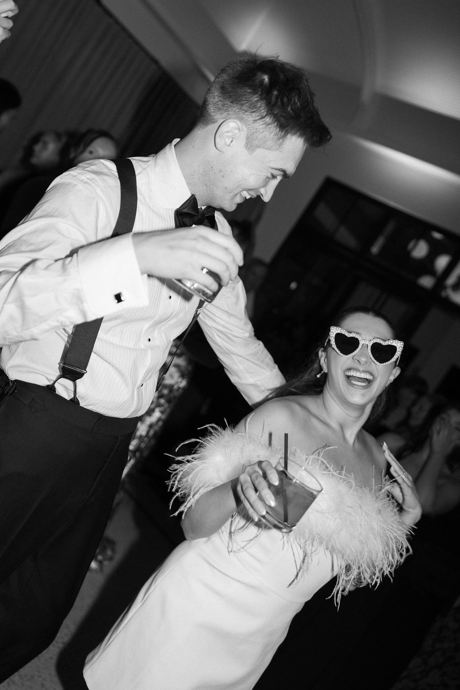 Black and white photo of a laughing bride in heart sunglasses and feather dress dancing with groom in suspenders at wedding reception.