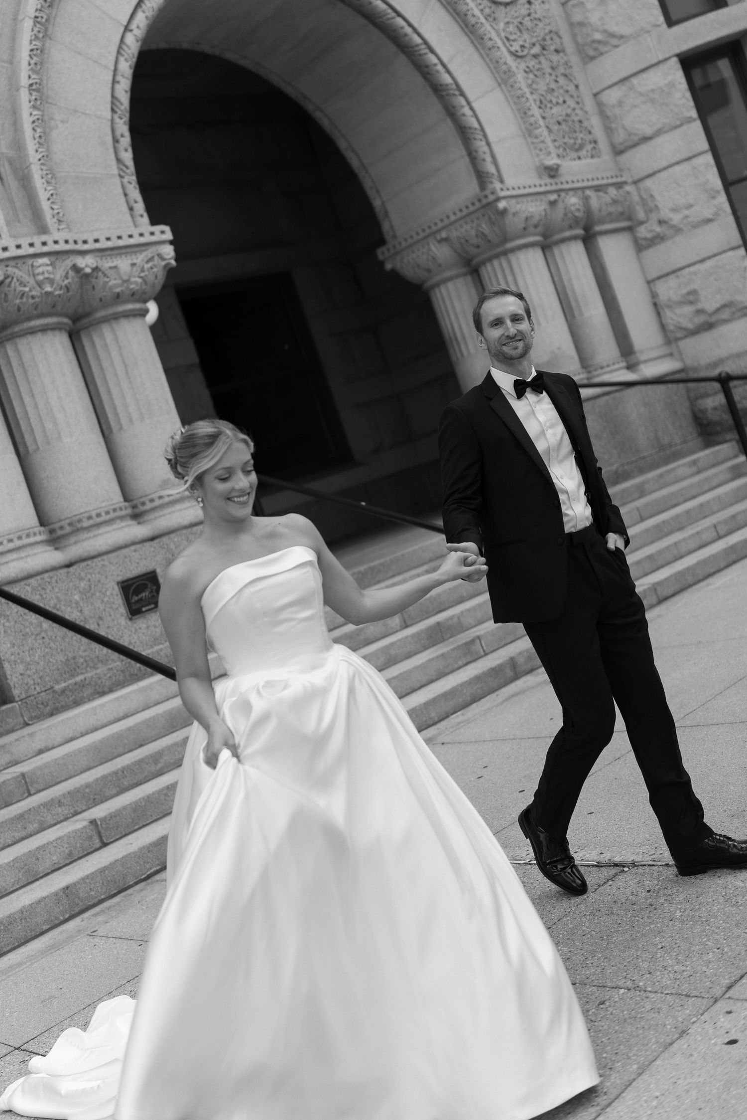 A bride in a white ballgown and groom in black tuxedo pose together in front of a stone building with arches.