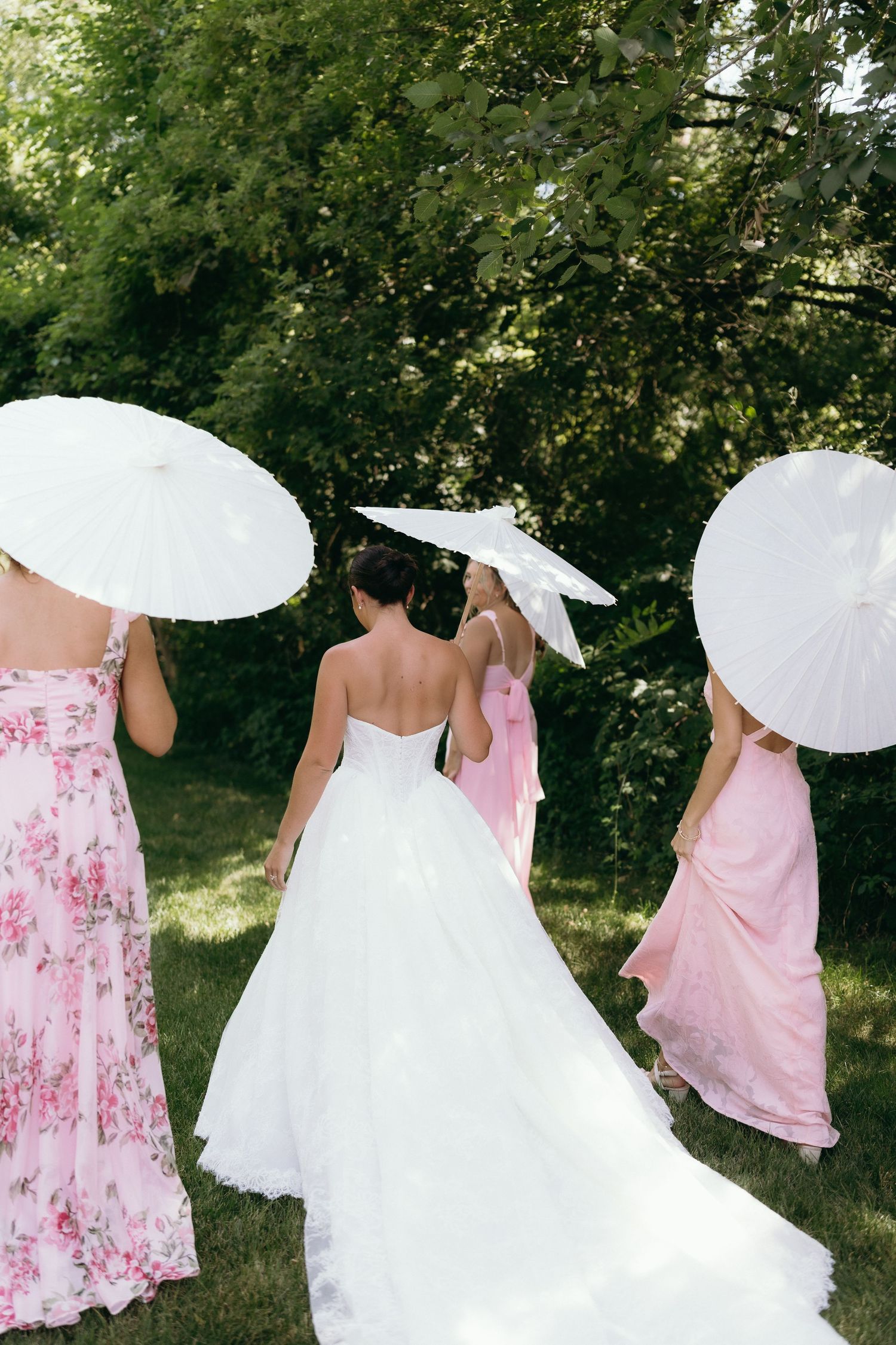 Bride in white gown walks with bridesmaids in pink dresses holding white parasols in a lush green garden.