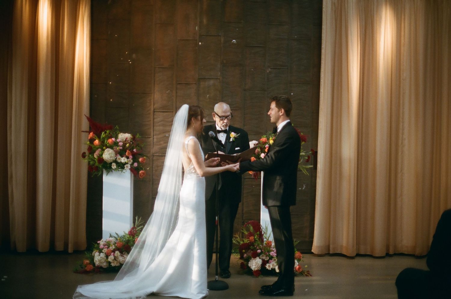 Bride and groom exchange vows at elegant indoor wedding ceremony with floral arrangements and draped curtains.