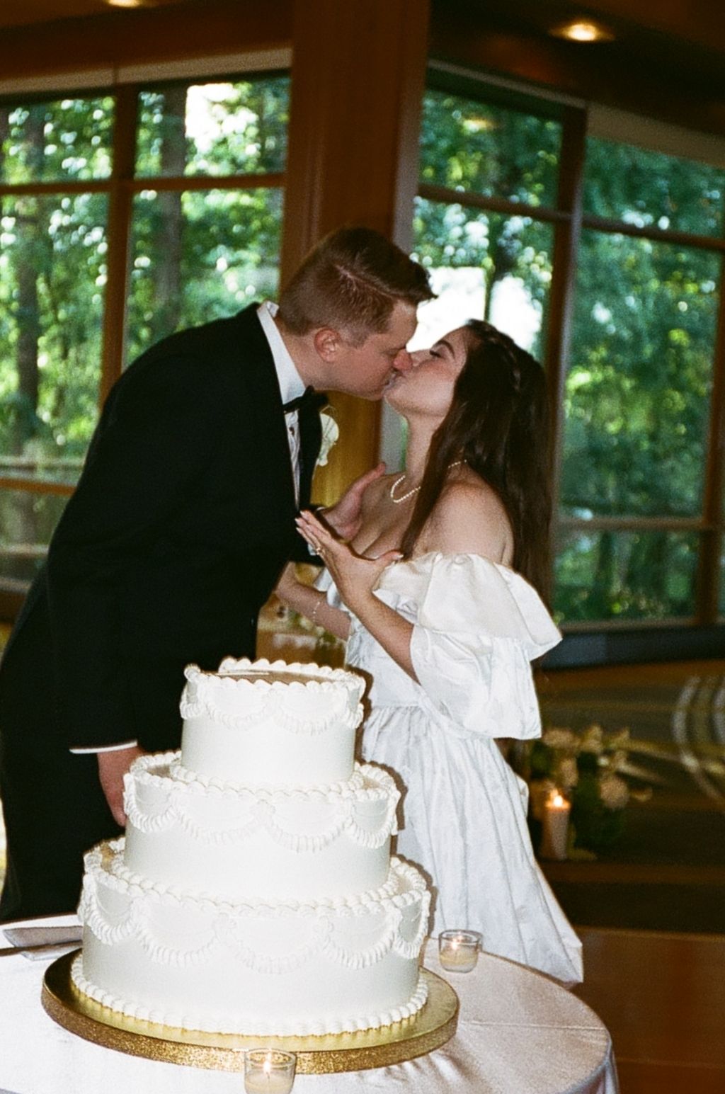 Bride and groom sharing a kiss while cutting their white three-tier wedding cake at their reception.