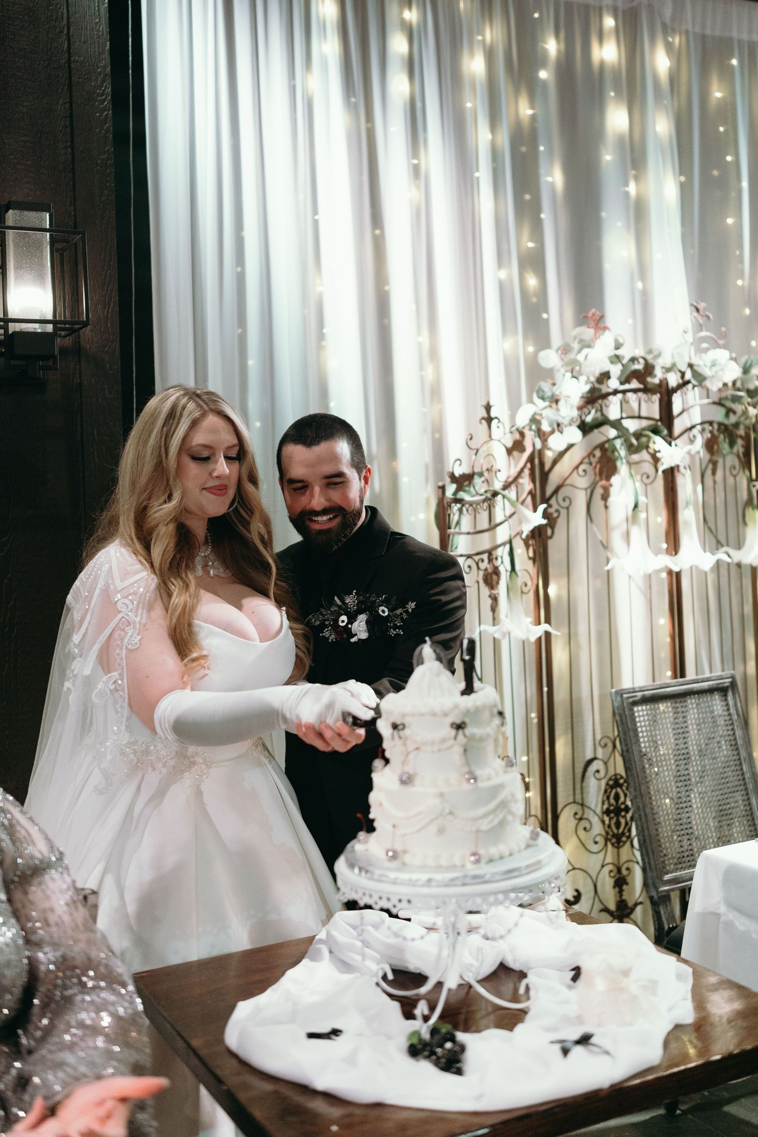 Couple cutting their elegant white tiered wedding cake under string lights.