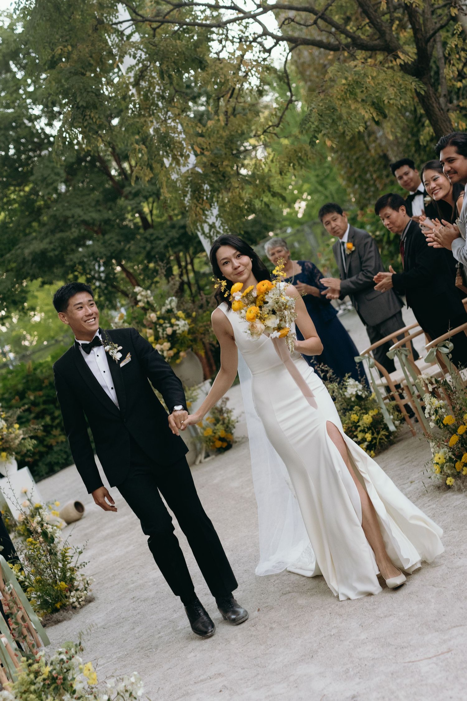 Newlyweds walk down aisle at outdoor garden ceremony with yellow floral accents.