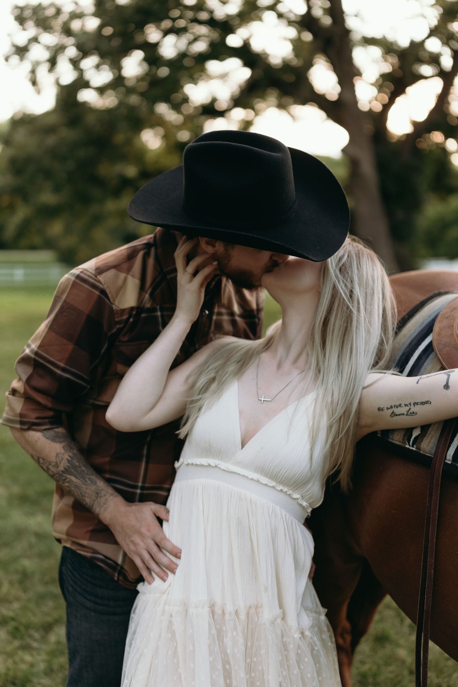 A romantic maternity photoshoot features a couple in western attire embracing next to a horse saddle at sunset.