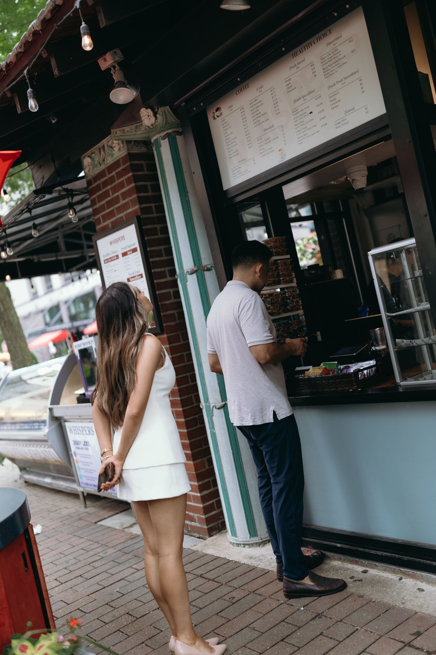 Customers line up at an exterior counter window of a brick building with menu boards displayed above.