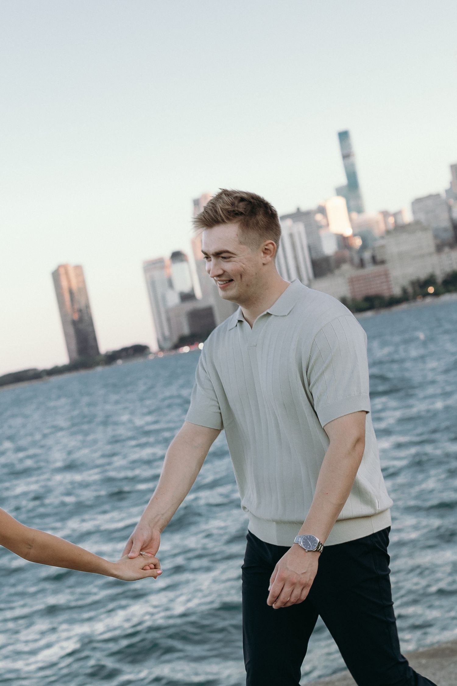 A person in grey polo and dark pants poses by the water with Chicago skyline in the background at sunset.