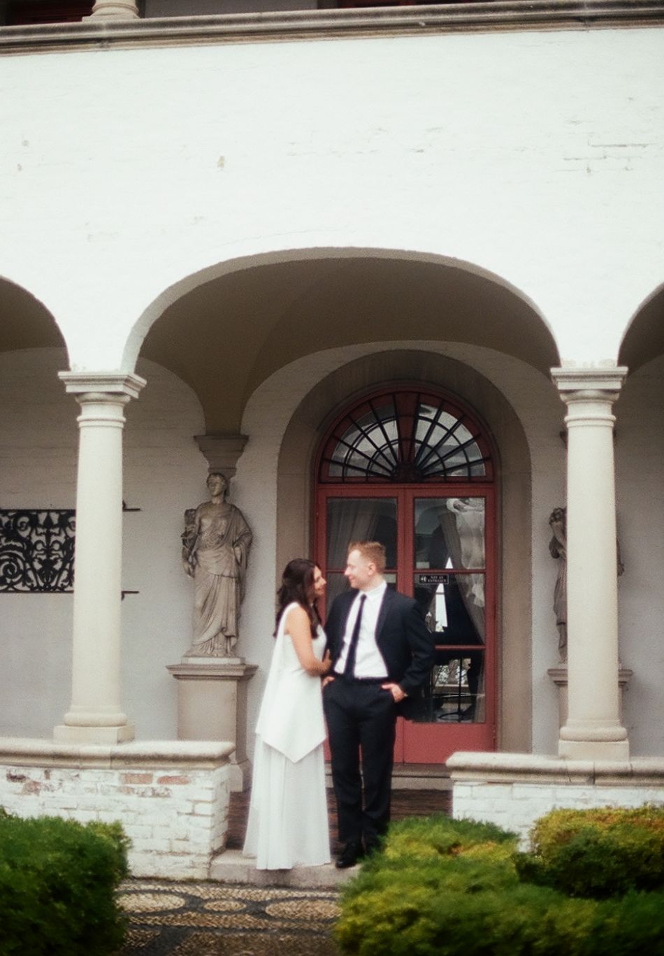 Couple standing together in a romantic outdoor colonnade with white columns and arched architectural details.