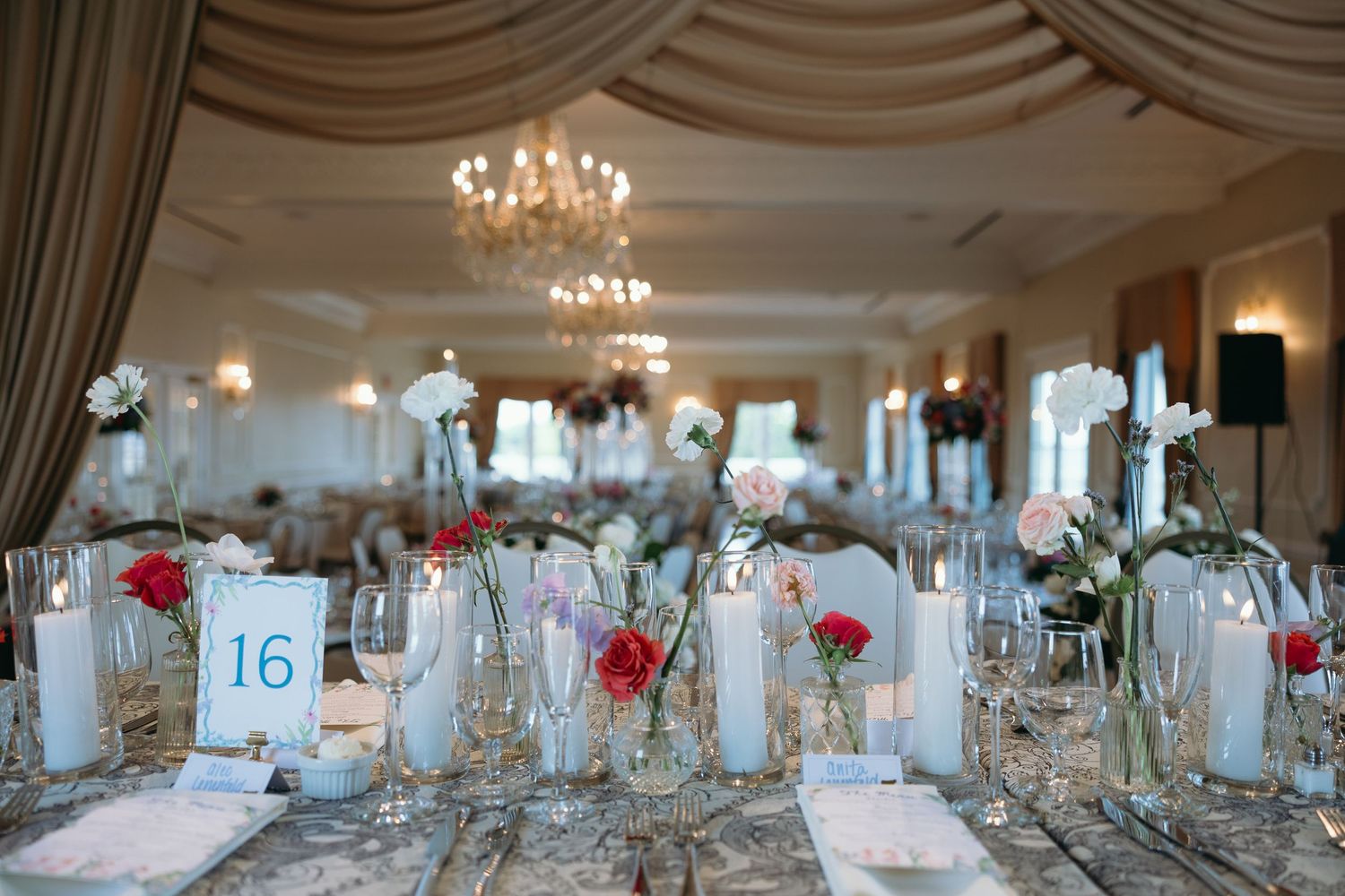 Elegant wedding reception venue with crystal chandeliers, draped white ceilings, and red floral centerpieces on tables.