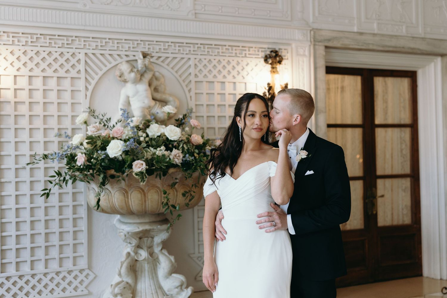 A wedding portrait set against a textured white wall with an ornate floral arrangement.