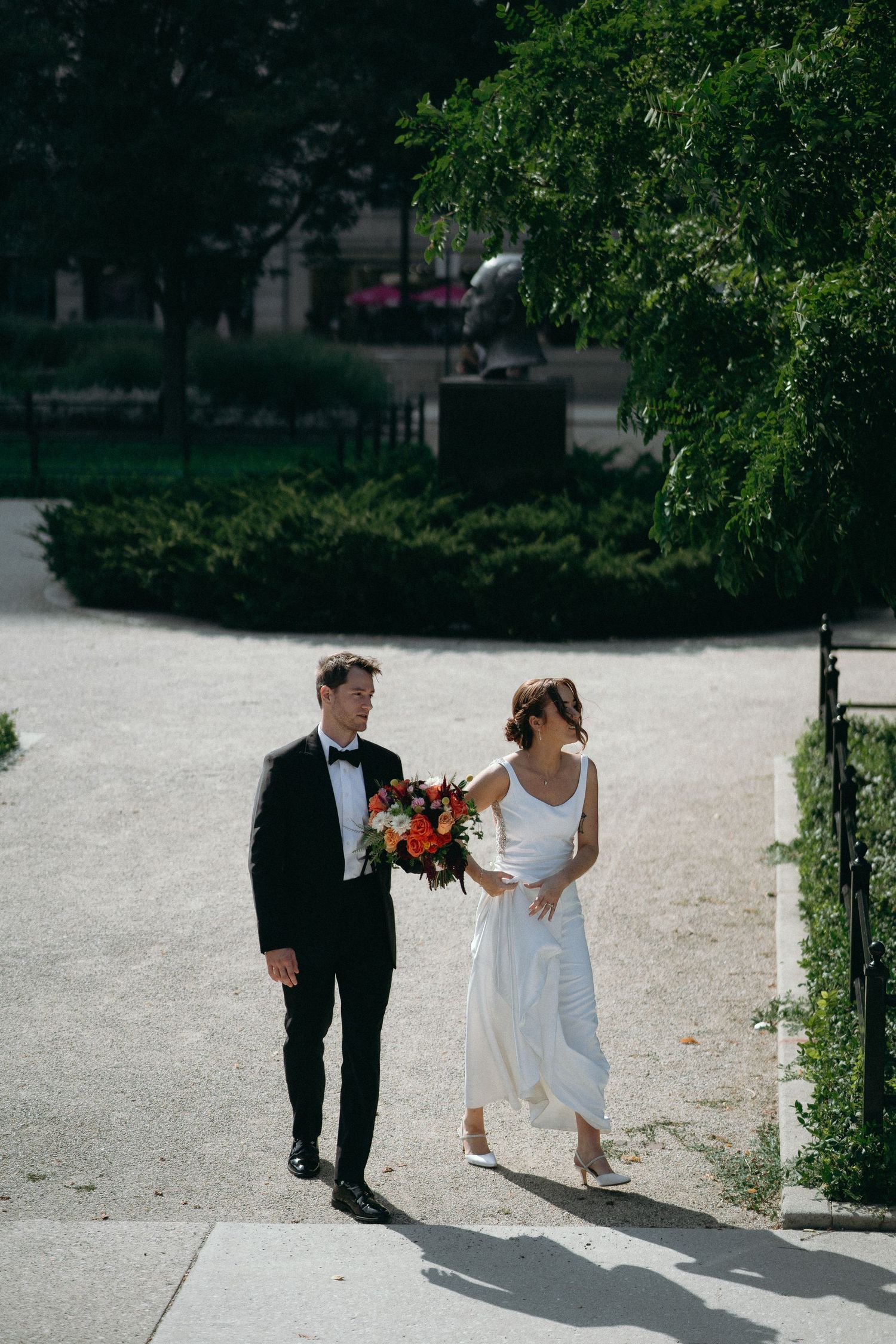 A couple in wedding attire walks along a sunlit path bordered by lush green trees and bushes.