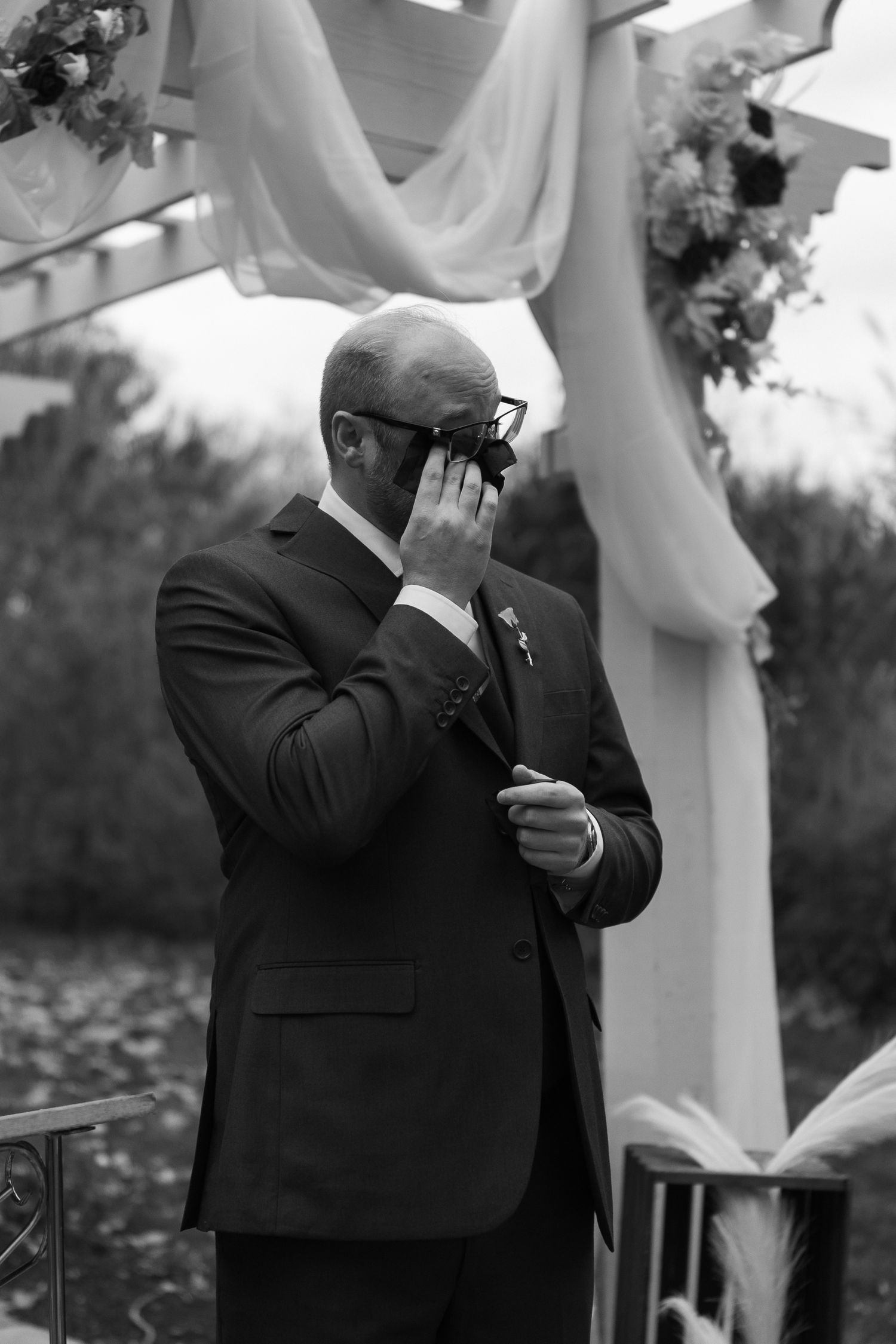 A groom becomes emotional and wipes away tears during an outdoor wedding ceremony under white fabric draping.