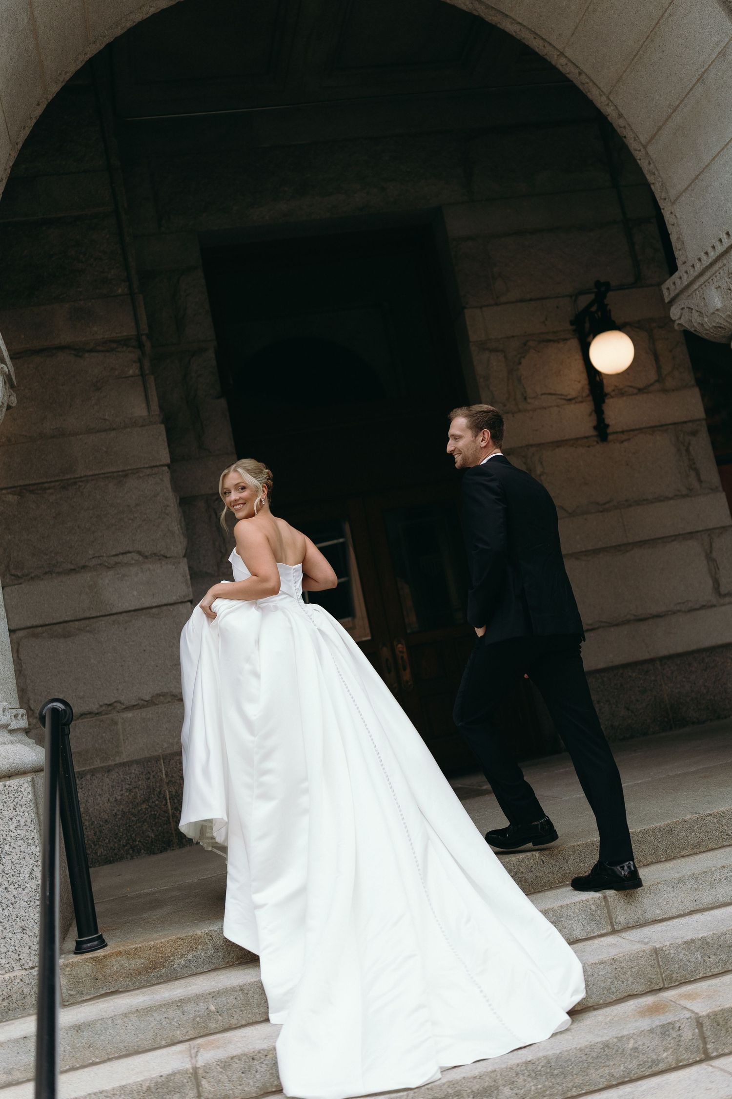 A bride in a flowing white wedding gown ascends stone steps while holding her dress train.