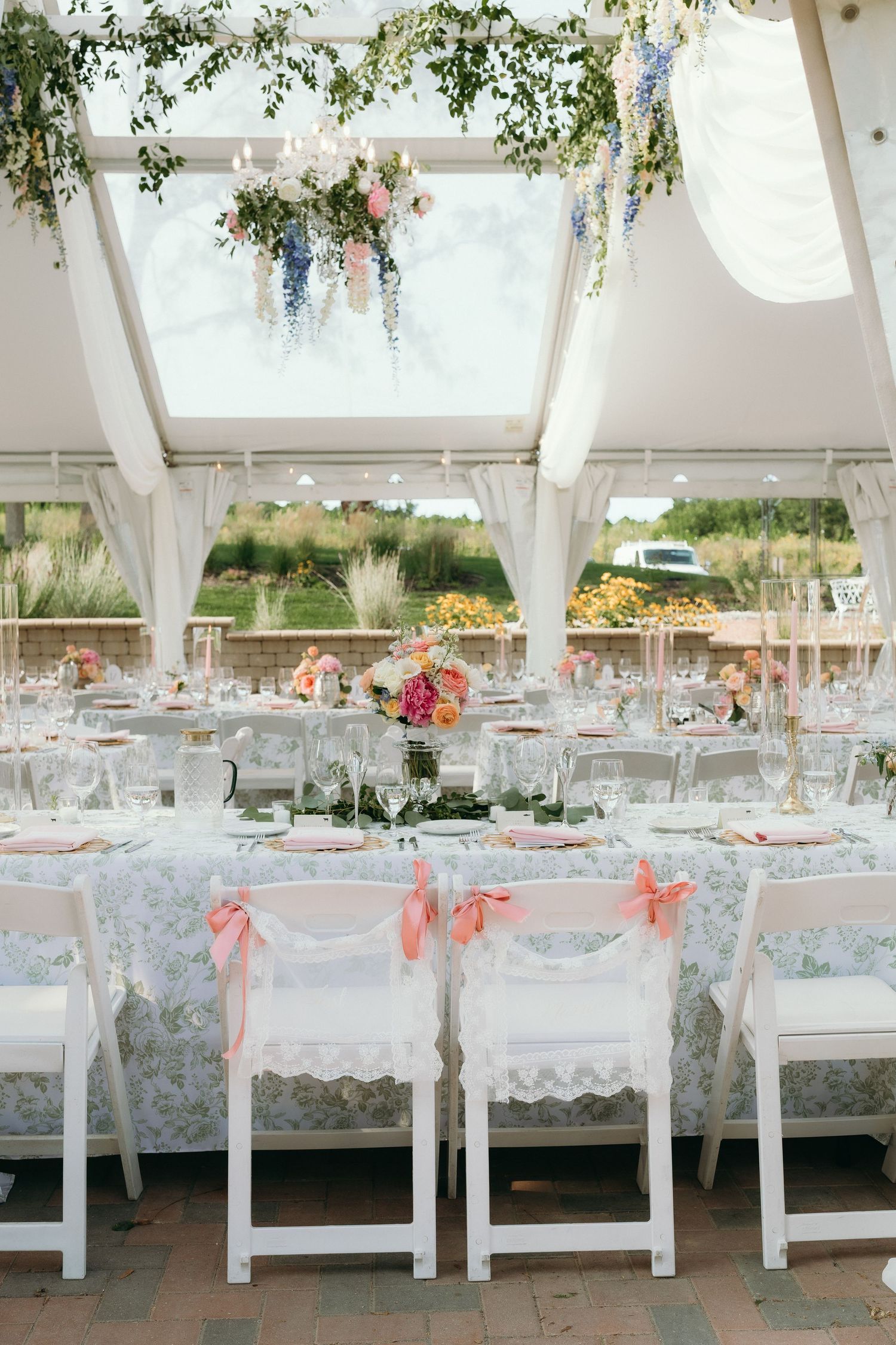 Bright wedding reception marquee with floral ceiling decor, blue floral tablecloths, and white chairs with pink bows.