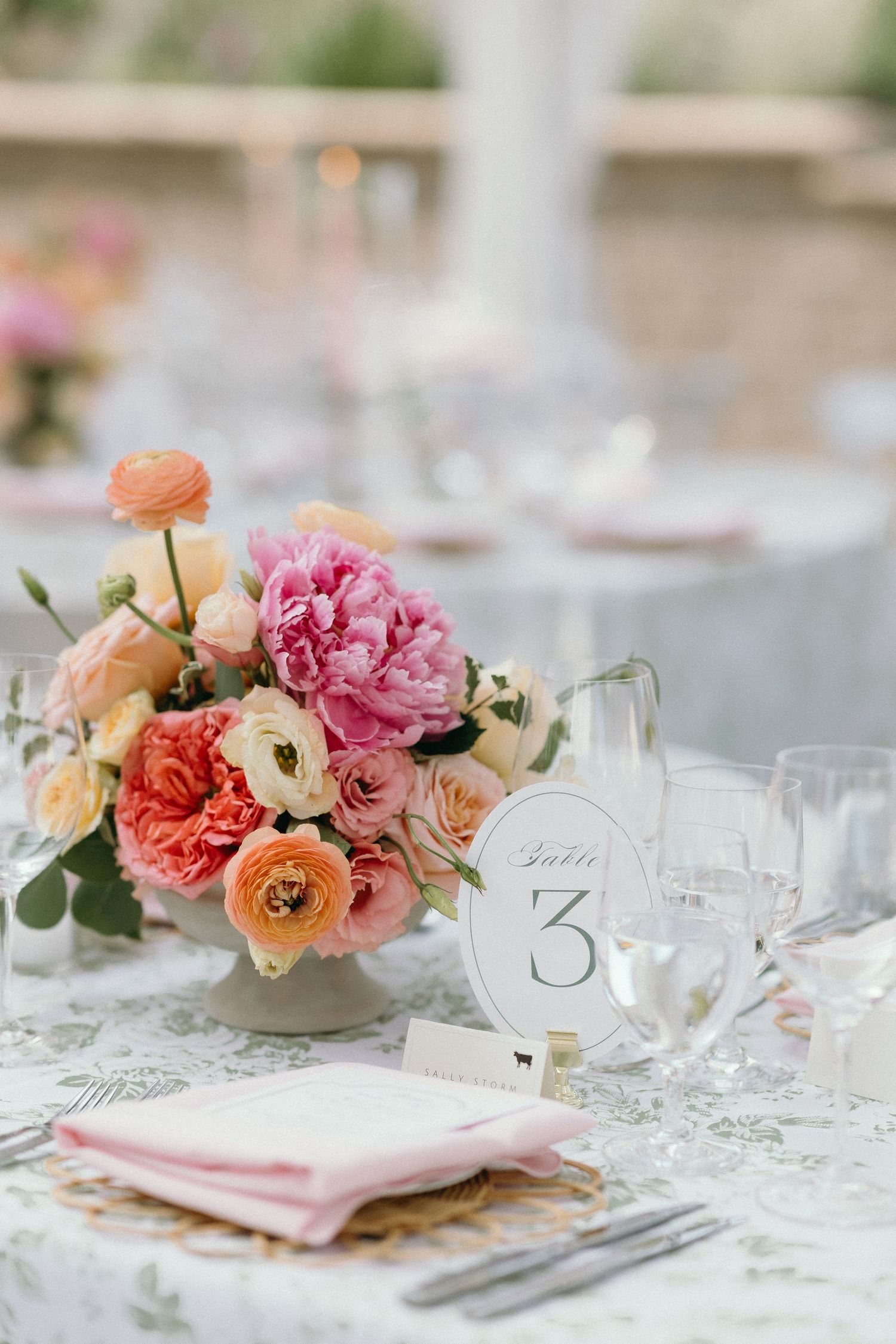 Elegant wedding table centerpiece with pink peonies, coral ranunculus, and peach roses beside a Table 3 sign.