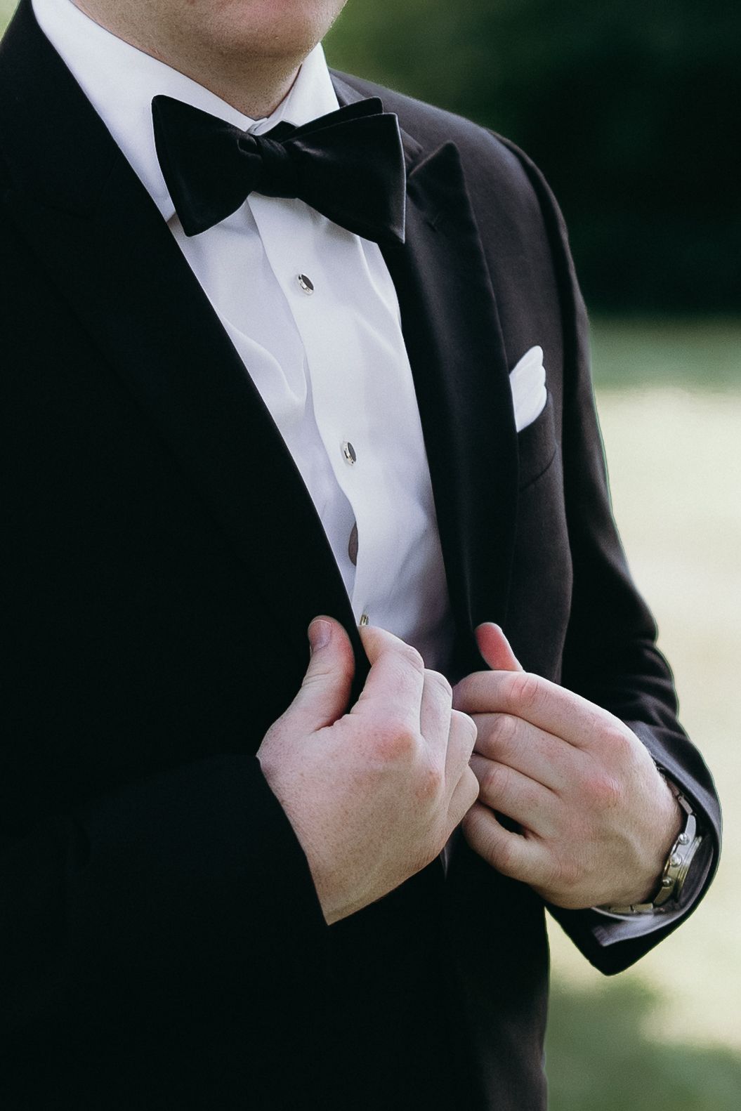 Man in black tuxedo with bow tie adjusting jacket lapels, wearing a watch and white pocket square.